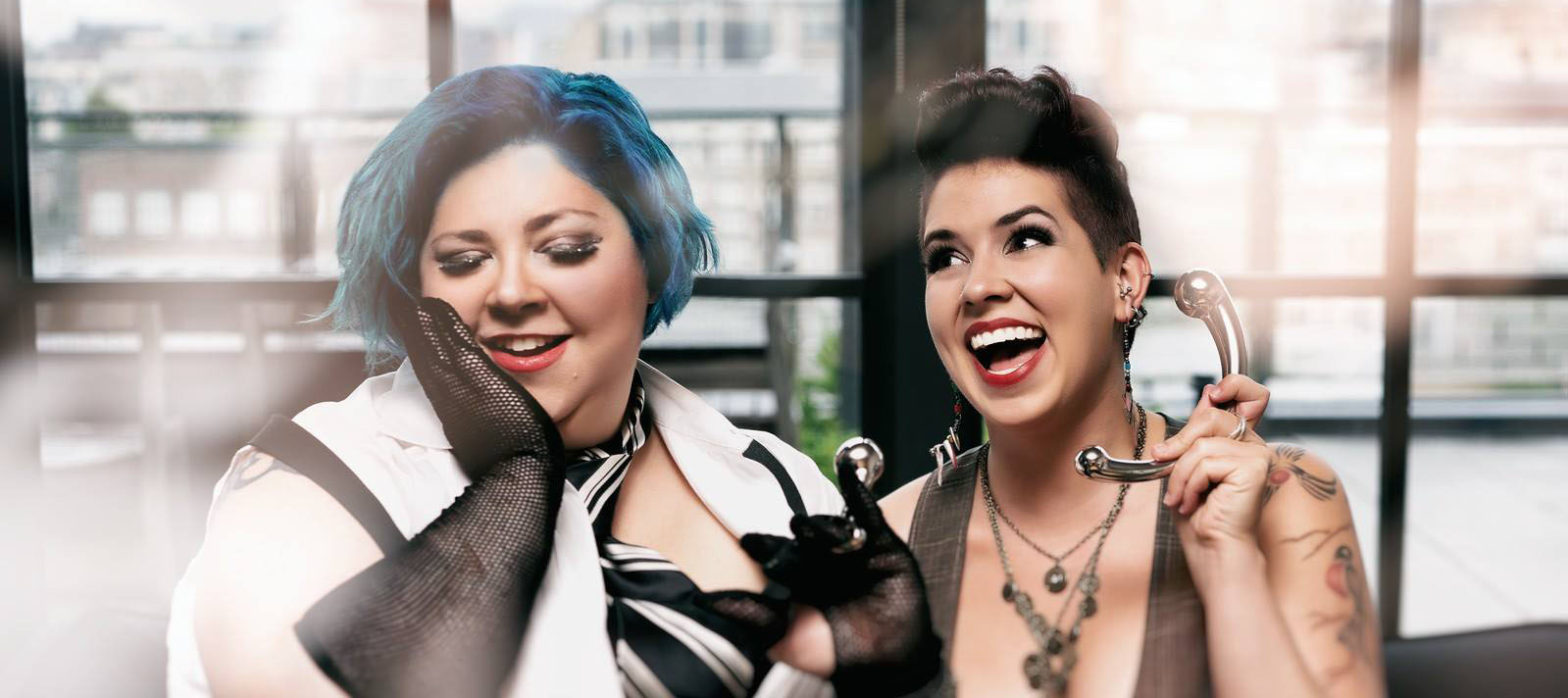 Image of Diva Darling wearing an Njoy butt plug like a ring and smiling, beside Mindi Mimosa holding an Njoy Pure Wand to her ear like a phone.