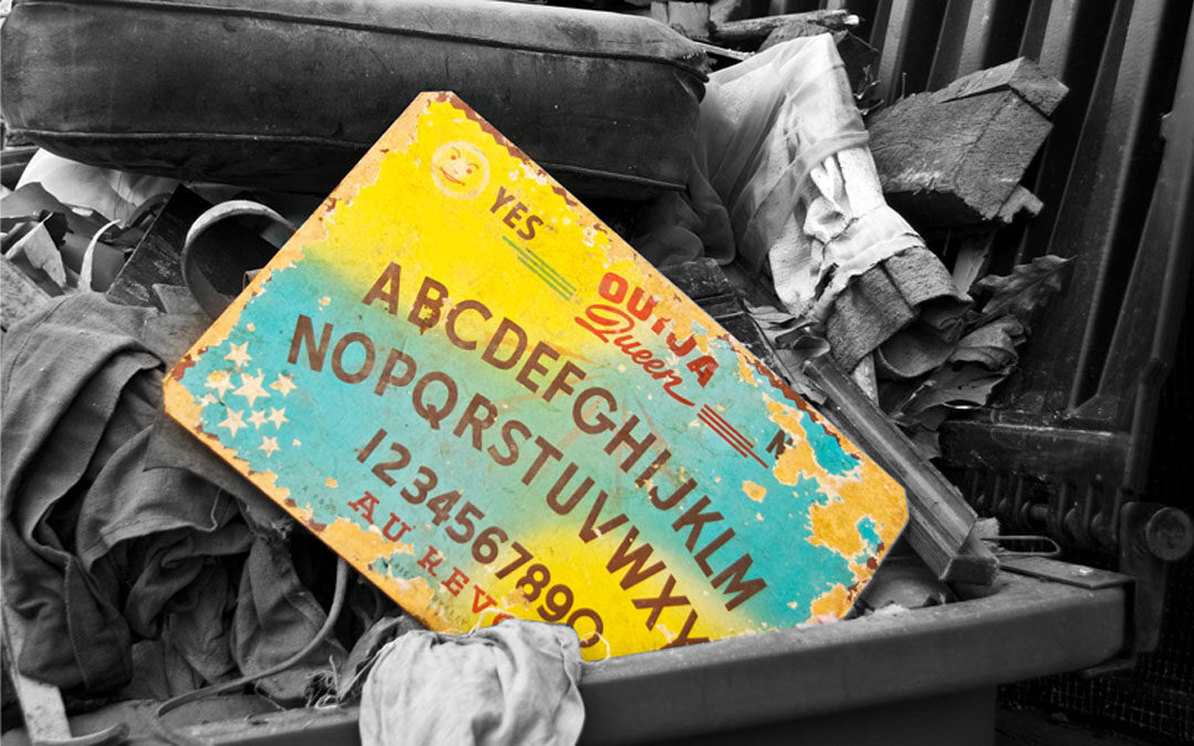 Image of a colorful "Ouija Queen" ouija board in a black-and-white dumpster full of trash